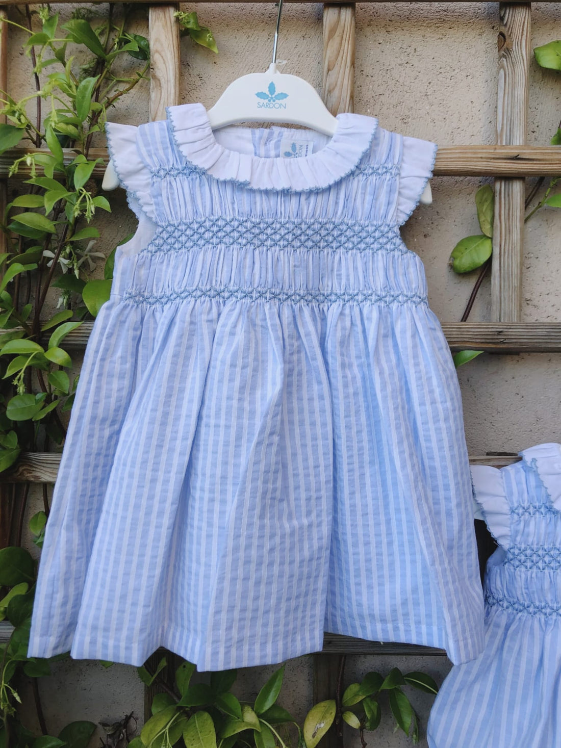 Vestido Agatha, en rayas celeste y blanco, sin mangas que van rematadas con volantito blanco con festón celeste al igual que el cuello. esta bordado en el delantero con punto smock también en celeste.