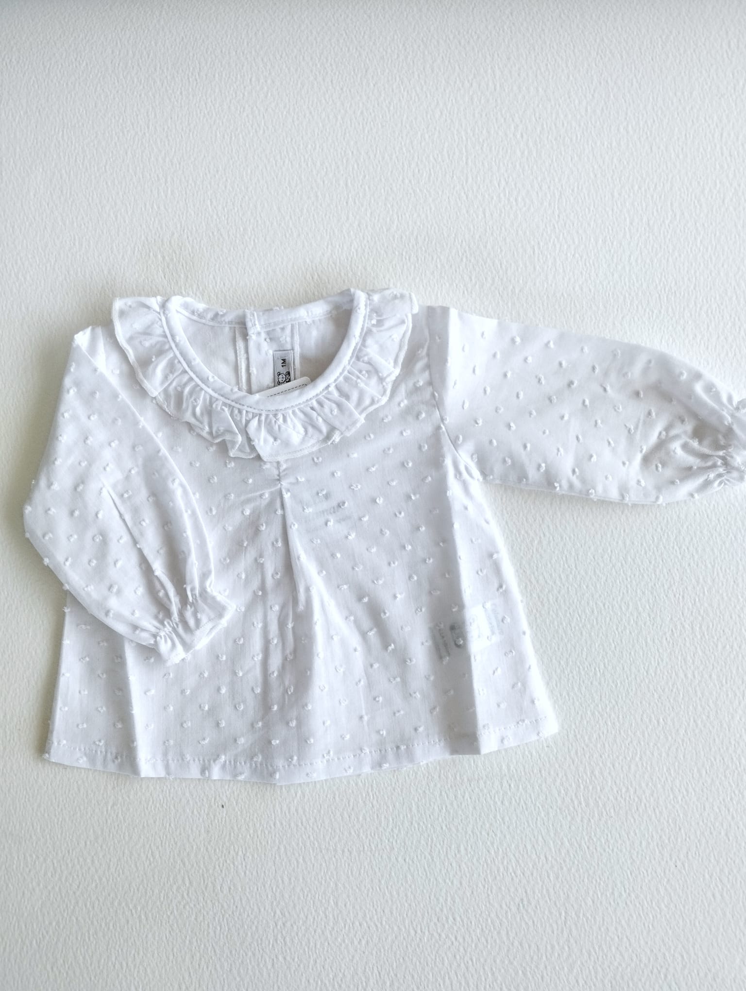Camisa plumeti manga larda con cuello de volante y puños funcidos, en color blanco va abotonada en la espalda.