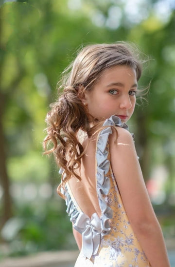 Bañador para niña estampado con flores y pájaros en blanco y gris con fondo amarillo, va ribeteado con volante gris y espalda en pico rematada con un lazo